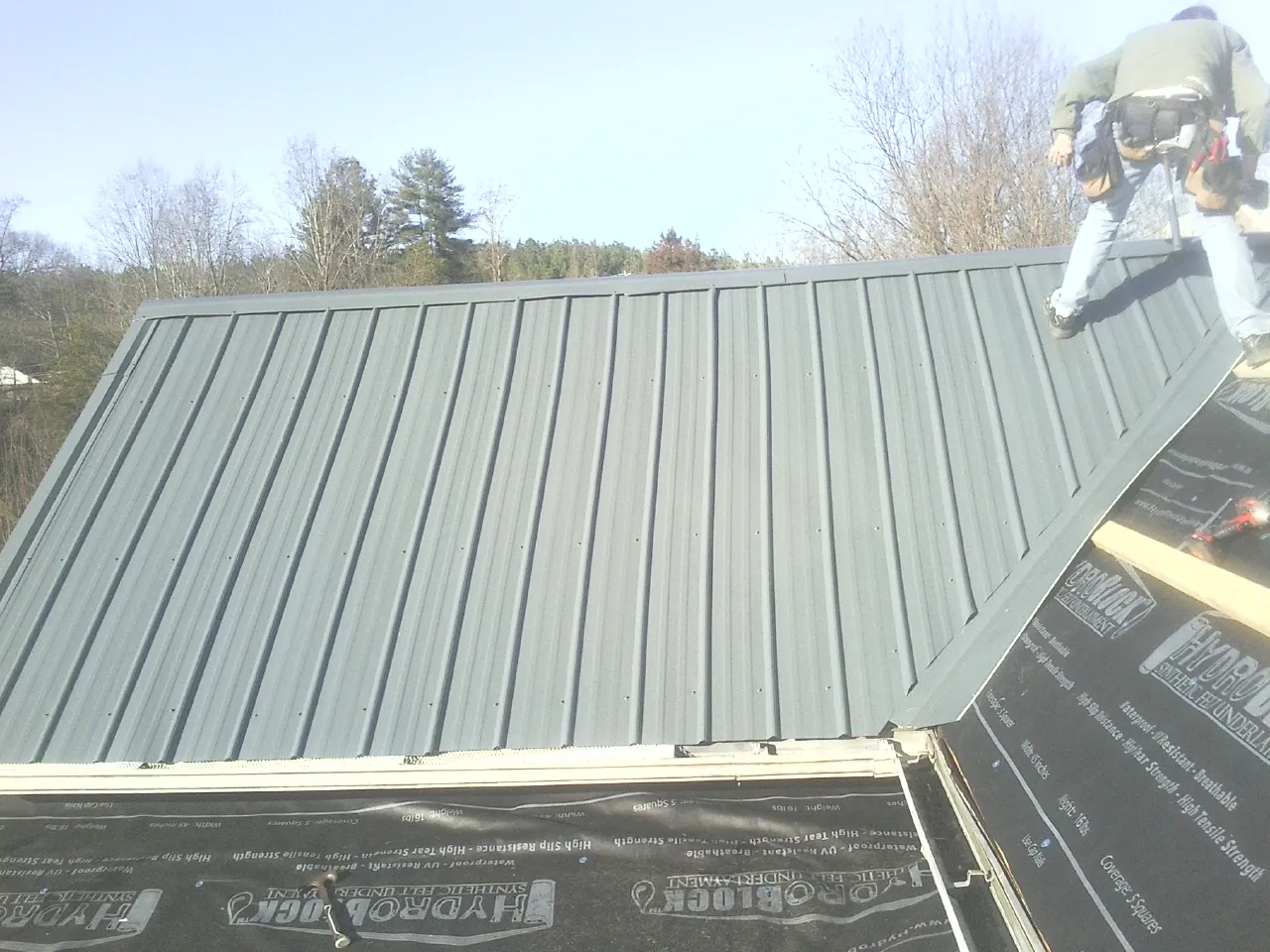 Crew installing dark green standing seam panels over synthetic underlayment in Pedro Bay