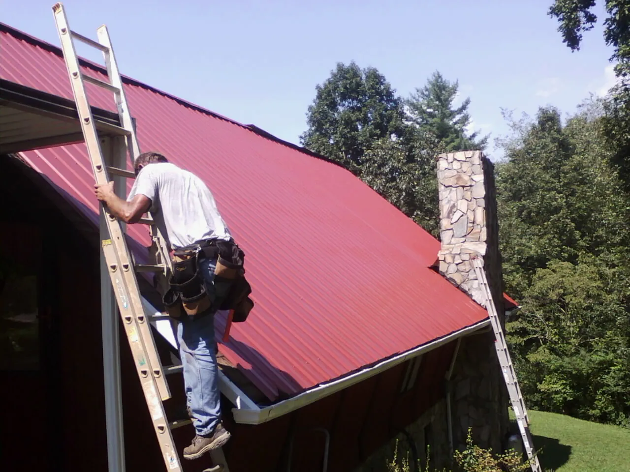Roofer on a ladder beside a red metal roof and stone chimney — Soffit & Fascia Repair by Roofing Brothers in Pedro Bay